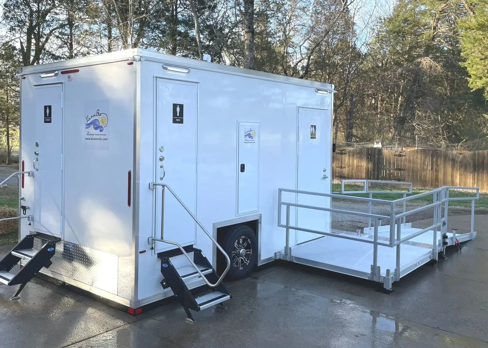 Mobile restroom trailer with ramp and steps. White exterior, black steps, metal ramp, parked outdoors.