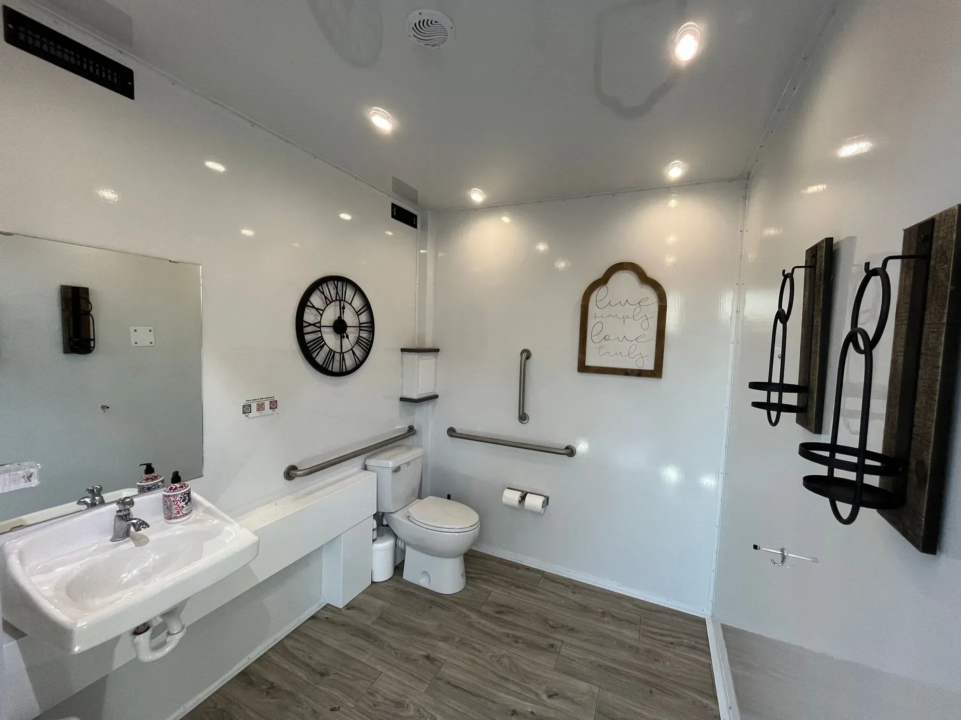 Accessible public restroom with sink, toilet, grab bars, clock, and decorative wall hangings.