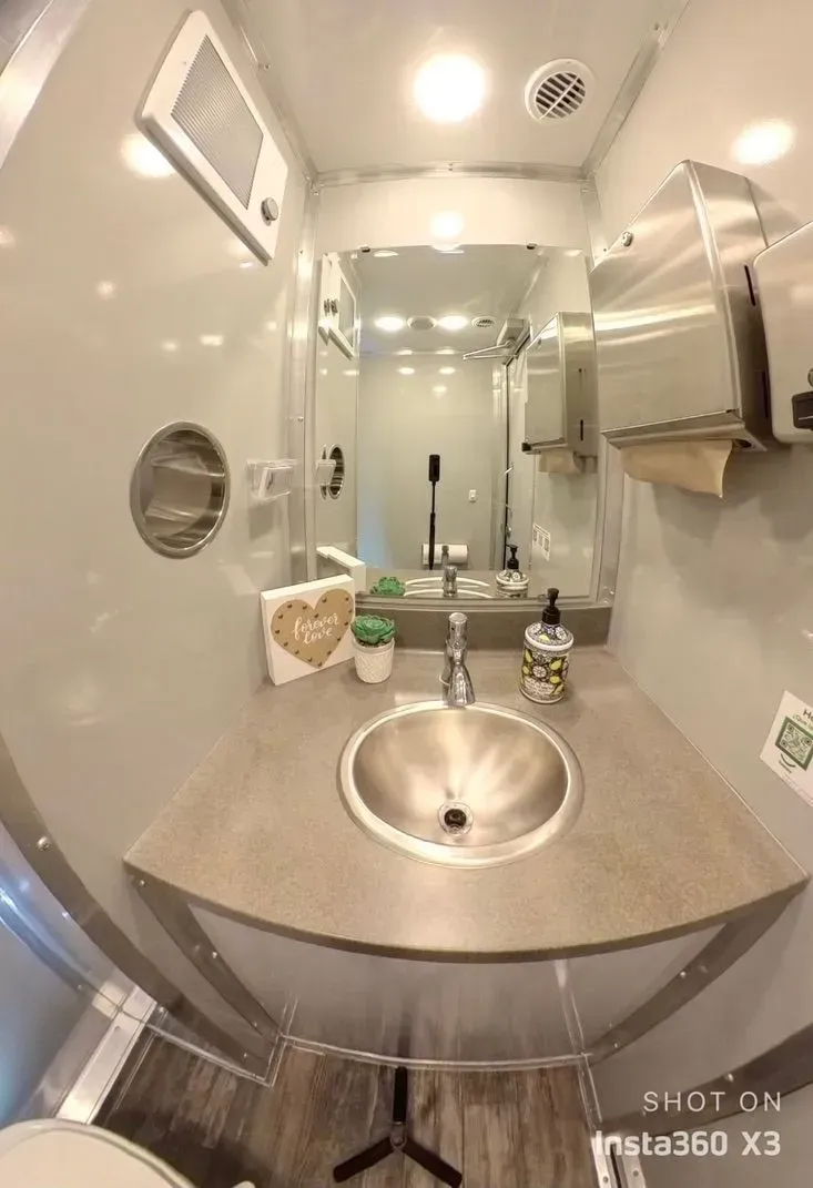 A well-lit, stainless-steel bathroom with a sink, mirror, and paper towel dispenser.