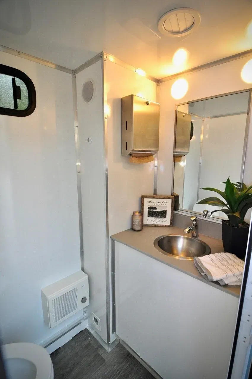 Interior view of a modern portable restroom. White walls, sink, mirror, and toilet visible.