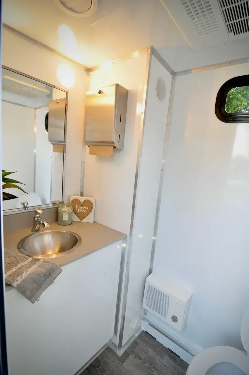 Interior of a luxury restroom trailer with a sink, mirror, and toilet. White walls and gray flooring.