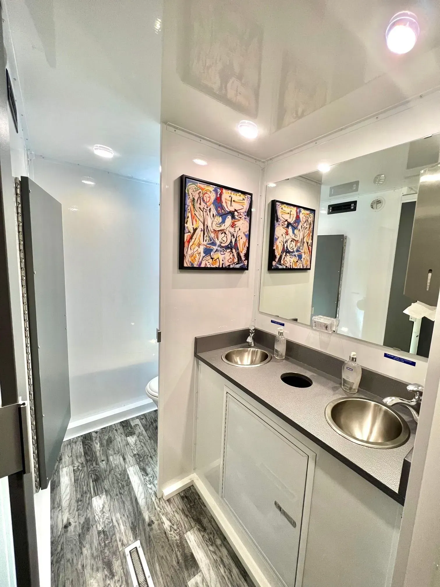 Interior of a portable restroom with two sinks, mirrors, and artwork on the wall.