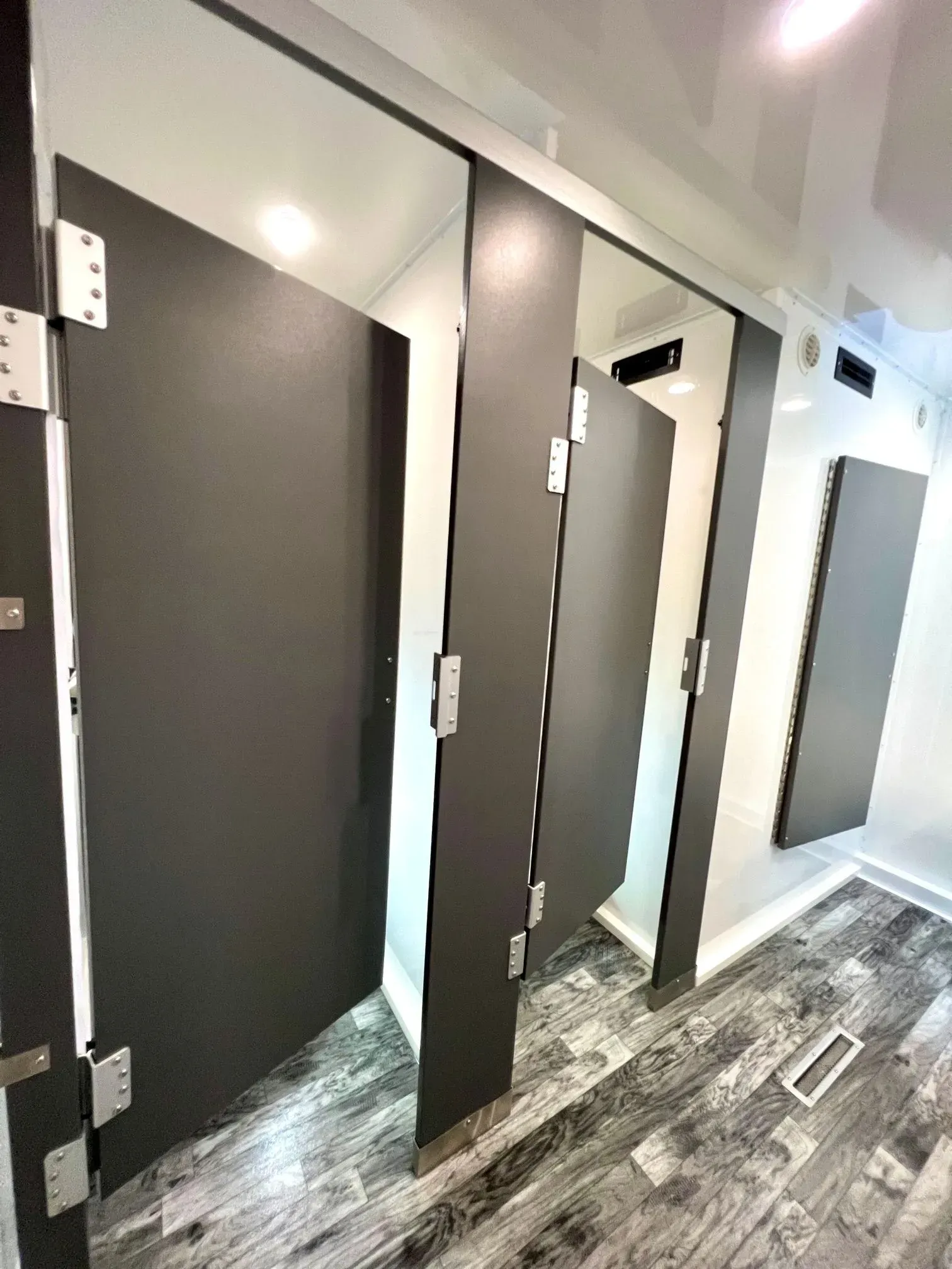 Restroom stalls with dark gray doors and silver hinges, set in a white and gray tiled space.