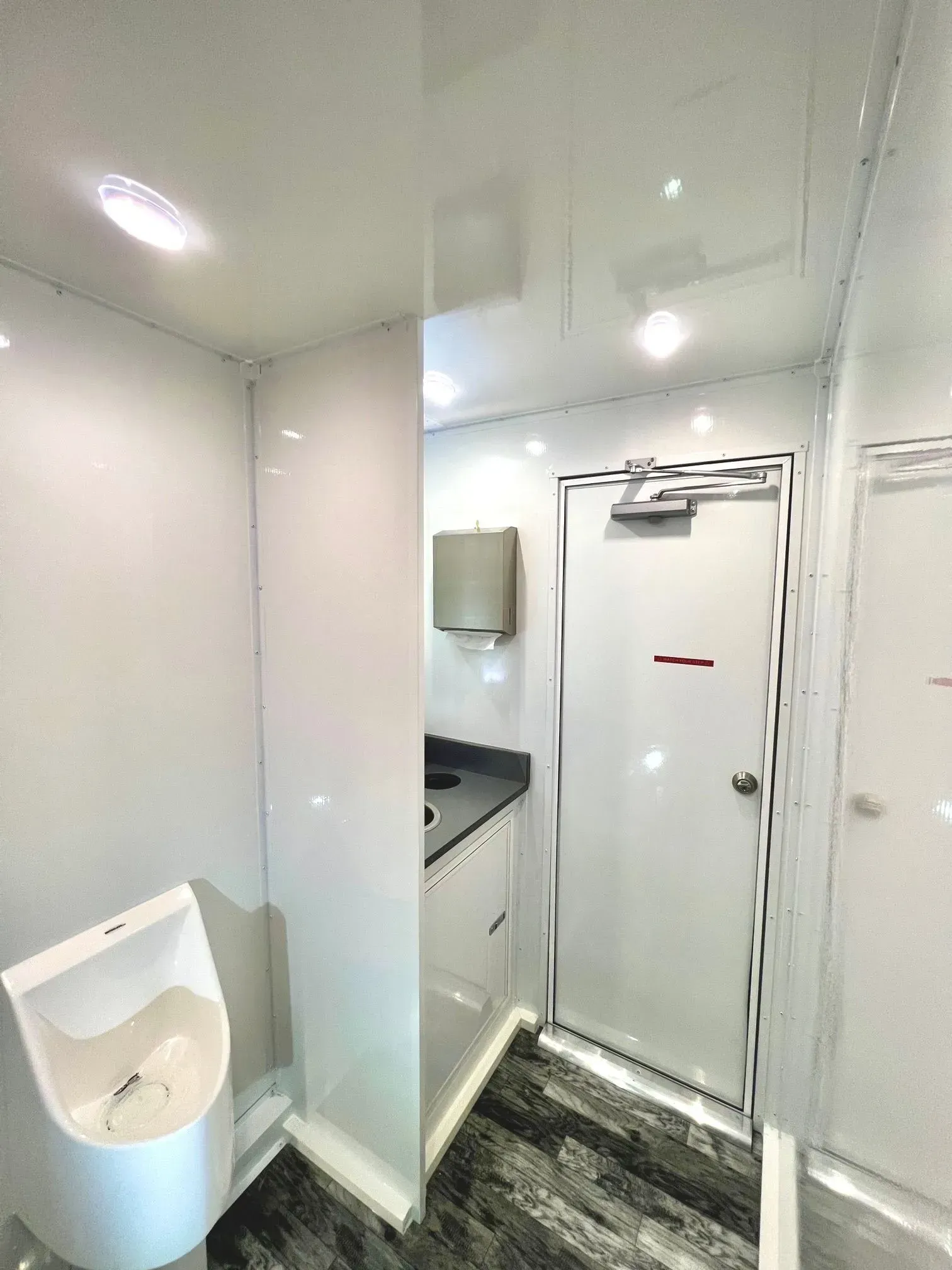 Interior of a clean, white portable restroom, featuring a urinal, sink, and door.