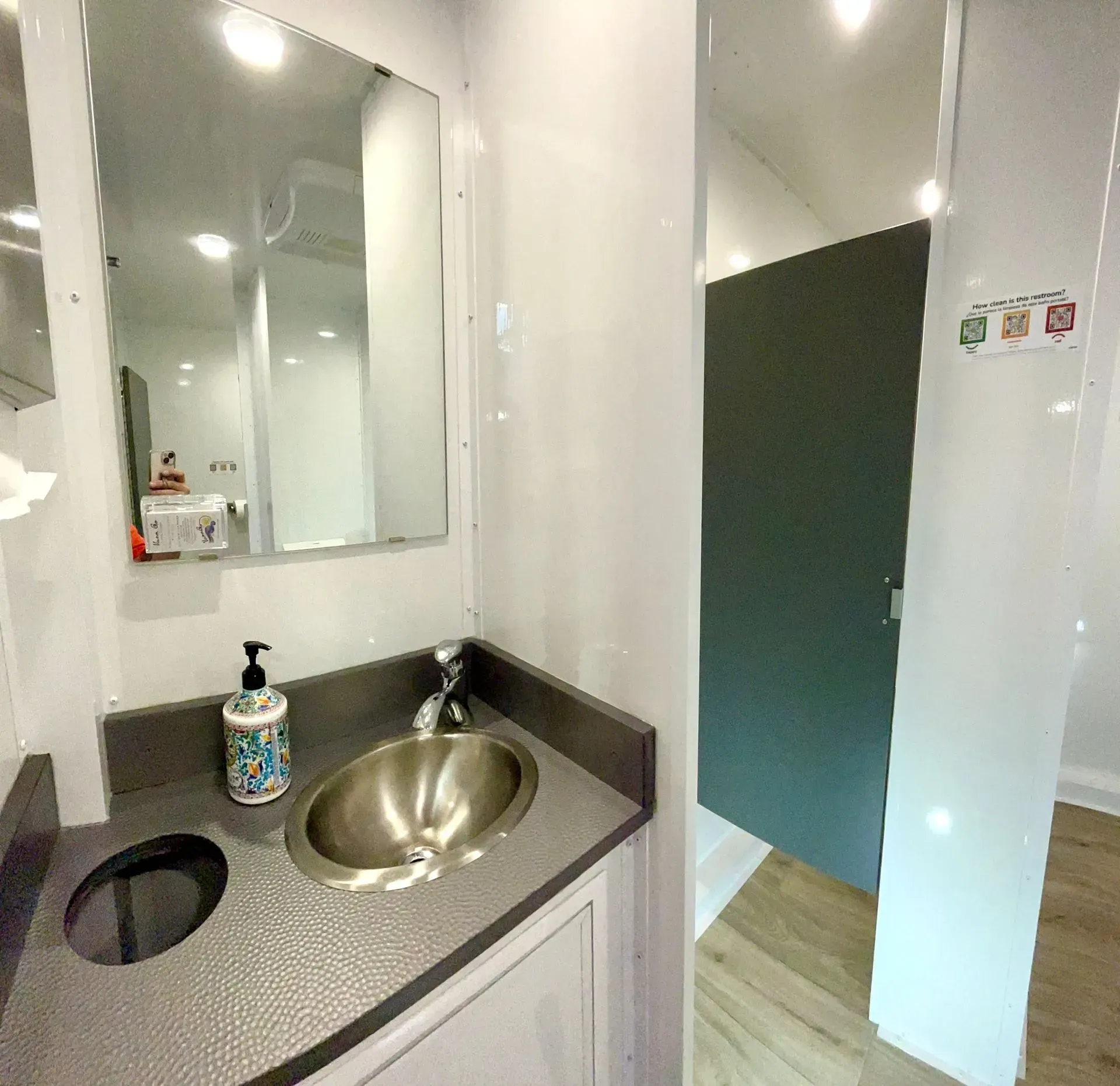 Interior of a portable restroom with a sink, mirror, and a stall door. Gray and white colors.