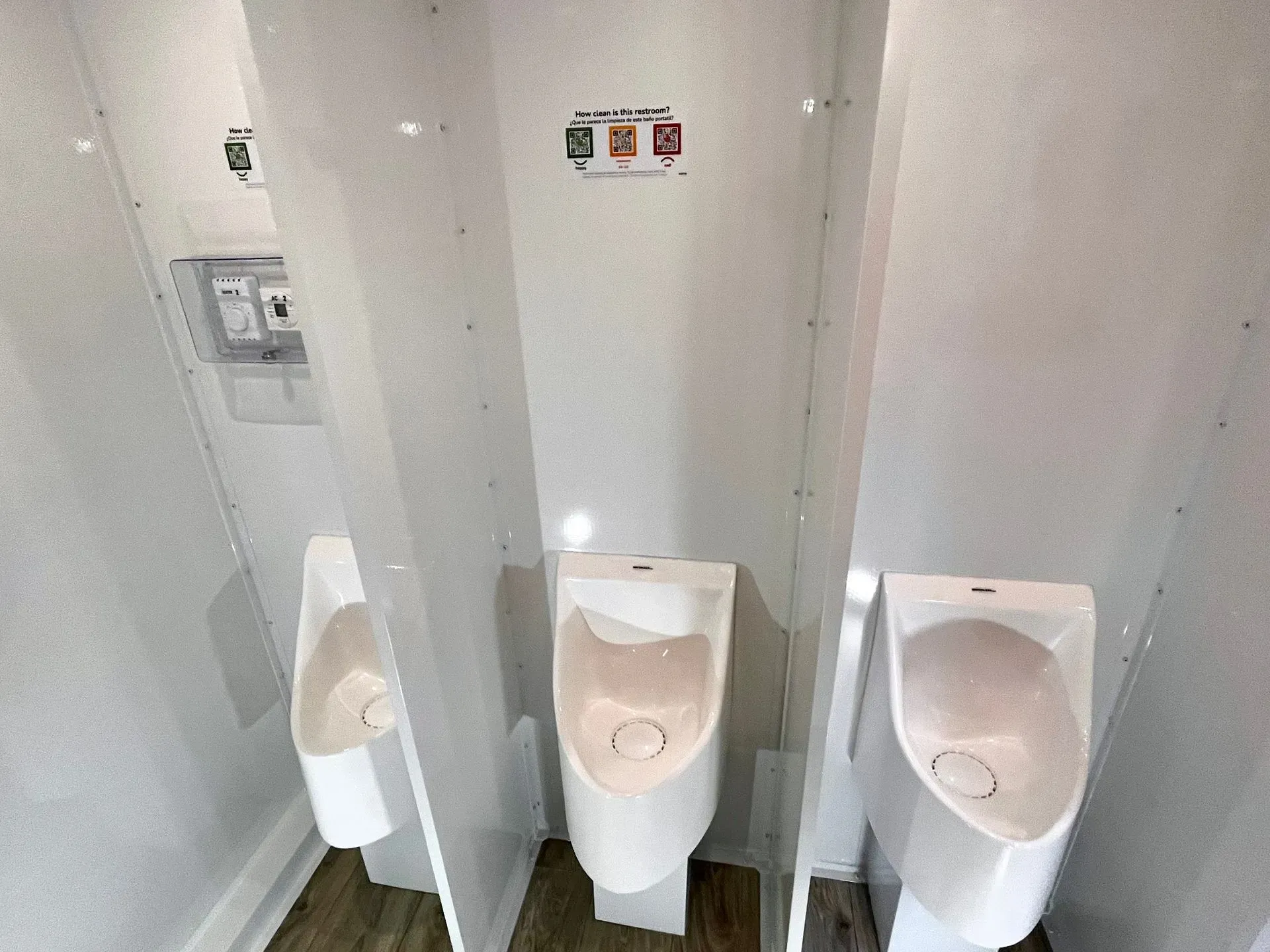 Three white urinals in stalls, wood-look floor, overhead lighting.
