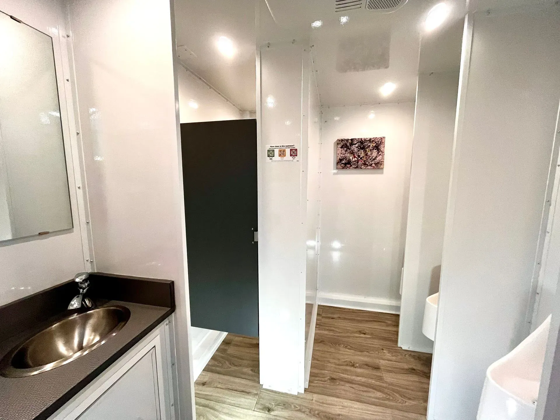 Interior of a portable restroom with a sink, mirror, and stalls. White walls, wood-look floor, overhead lights.