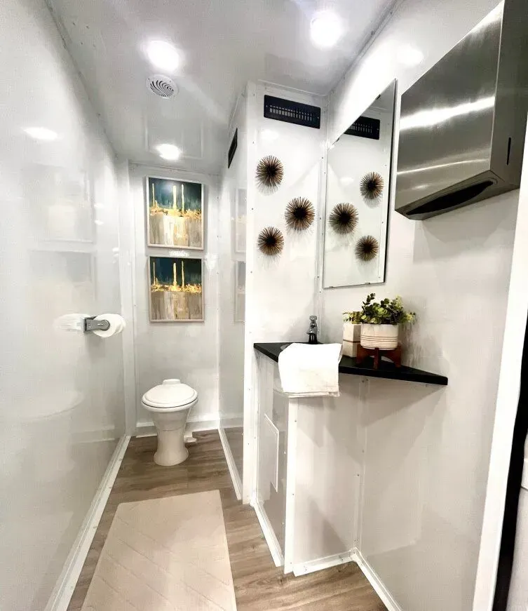 Modern, clean restroom trailer interior with toilet, sink, mirror, and decorative art. White walls, wood-look floor.