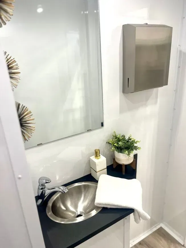 Bathroom with a stainless steel sink, mirror, and paper towel dispenser. A towel, soap dispenser, and plant are on the counter.