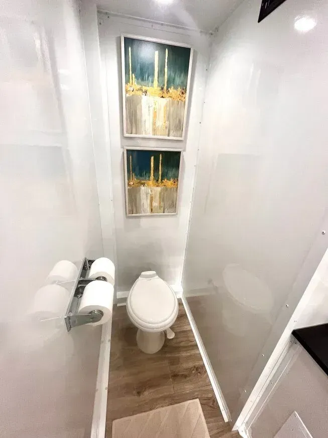 Interior of a portable restroom with white walls, art, a toilet, and tissue holder. Light wood-look flooring.