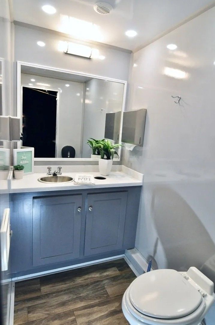 Interior of a modern portable restroom. Features a sink, mirror, toilet, and gray cabinets.