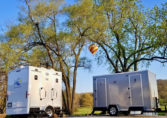 Two portable restroom trailers next to trees, a hot air balloon is in the distance.