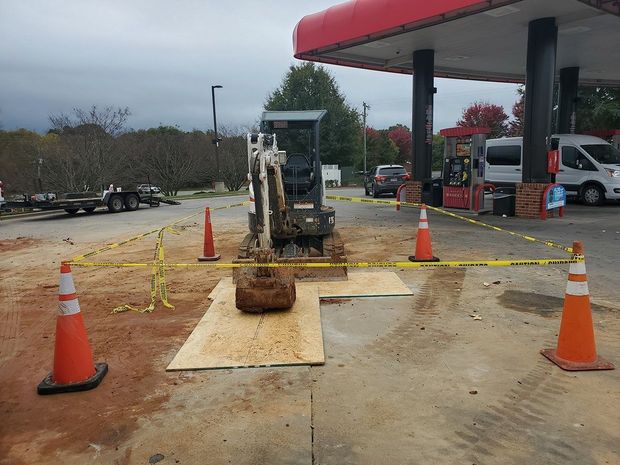 Mini excavator behind safety tape and cones at a gas station, possibly doing repair work.