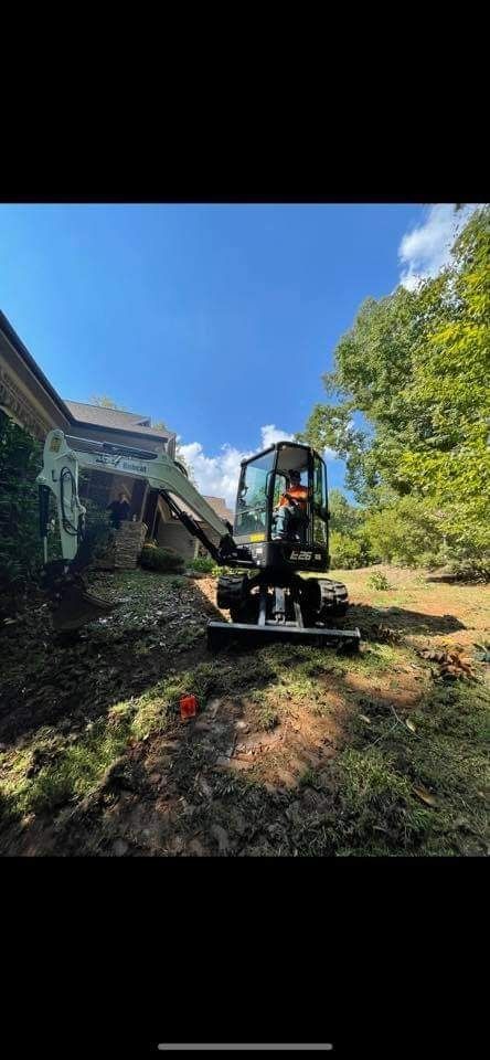 An excavator removing earth near a building under a blue sky, with trees on the right.