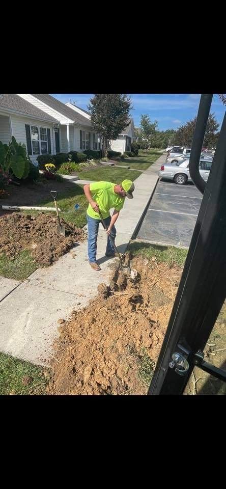 A person in a lime-green shirt digging with a shovel next to a sidewalk near a building.