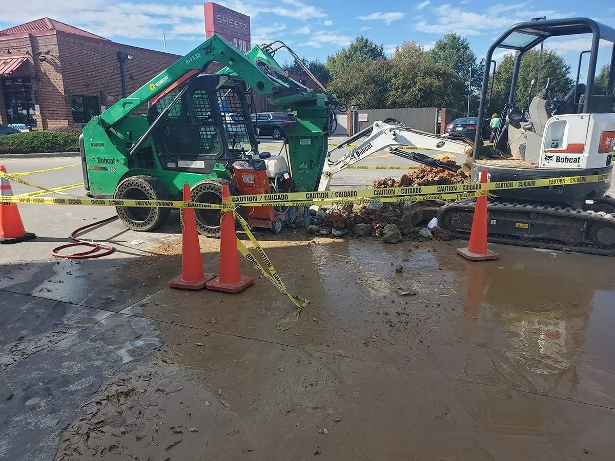Construction site with two machines and orange cones cordoned off with yellow tape; pavement is wet and muddy.