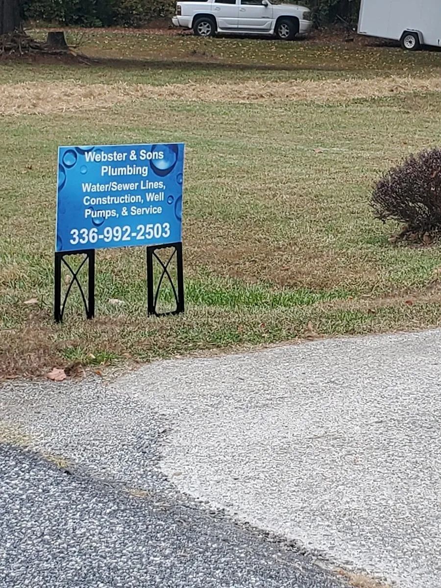 Sign for Webster & Son Plumbing in a grassy yard, with a phone number.