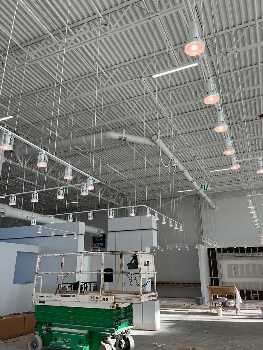Interior of a building under construction, showing overhead lighting fixtures, ductwork, and a scissor lift.