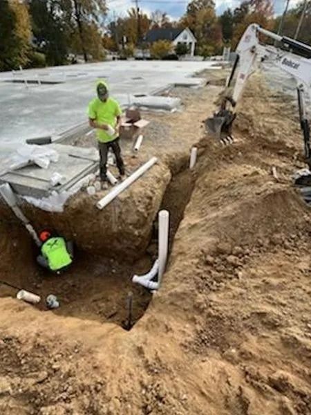 Construction site with workers, machinery, and trenches. Workers installing pipes in an open trench, surrounded by dirt and concrete.