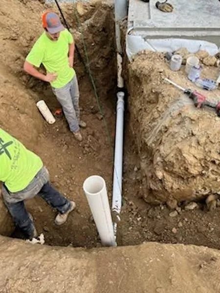 Two construction workers installing white PVC pipe in a trench. One stands, the other works.