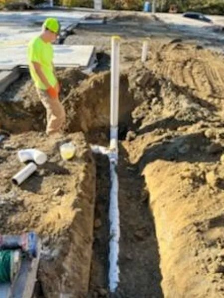 Construction worker near a trench with white pipes and a vertical vent pipe. Yellow safety vest and hard hat.