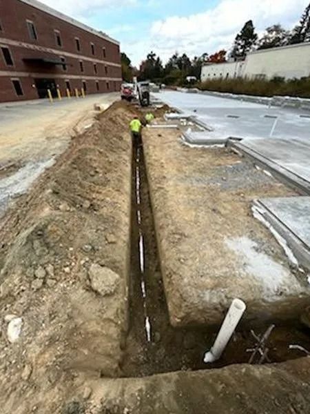 Construction workers in a long trench laying pipes next to a building and parking lot.