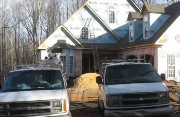 Two white vans parked in front of a house under construction; wood, windows, and scaffolding visible.