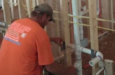 Plumber in orange shirt working on PVC pipes inside a wood-framed wall.