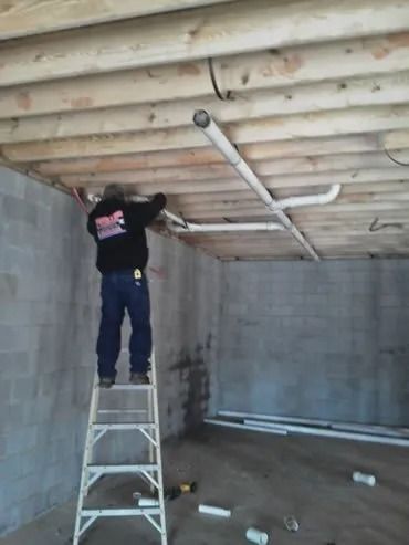 A person on a ladder installs PVC pipes on a ceiling in a concrete-walled room.