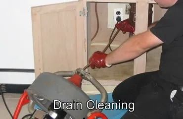 A person using a drain cleaning machine under a sink in a cabinet.