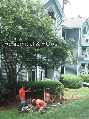 Workers installing a fence near a bush and apartment building, orange shirts, tools, sunny day.