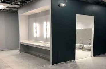 Interior of a partially constructed restroom with a sink area and toilet stalls. Walls are painted grey and dark teal.