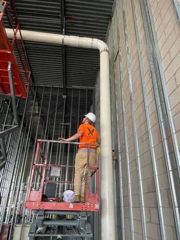 Construction worker in orange shirt on a lift, installing pipe against wall with metal studs.