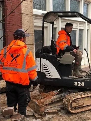 Two construction workers in orange safety vests; one operating a small excavator, the other observing outside a building.