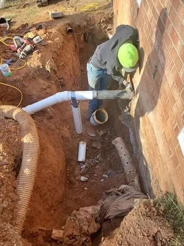 Person installing exterior drain pipe next to brick wall in a trench.