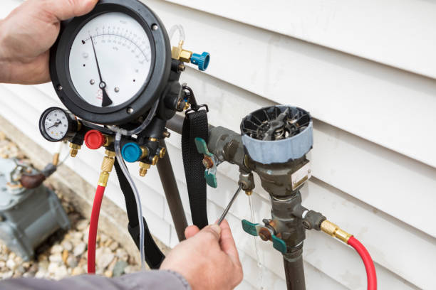 A technician uses a pressure gauge manifold to test a backflow prevention device on the side of a building.