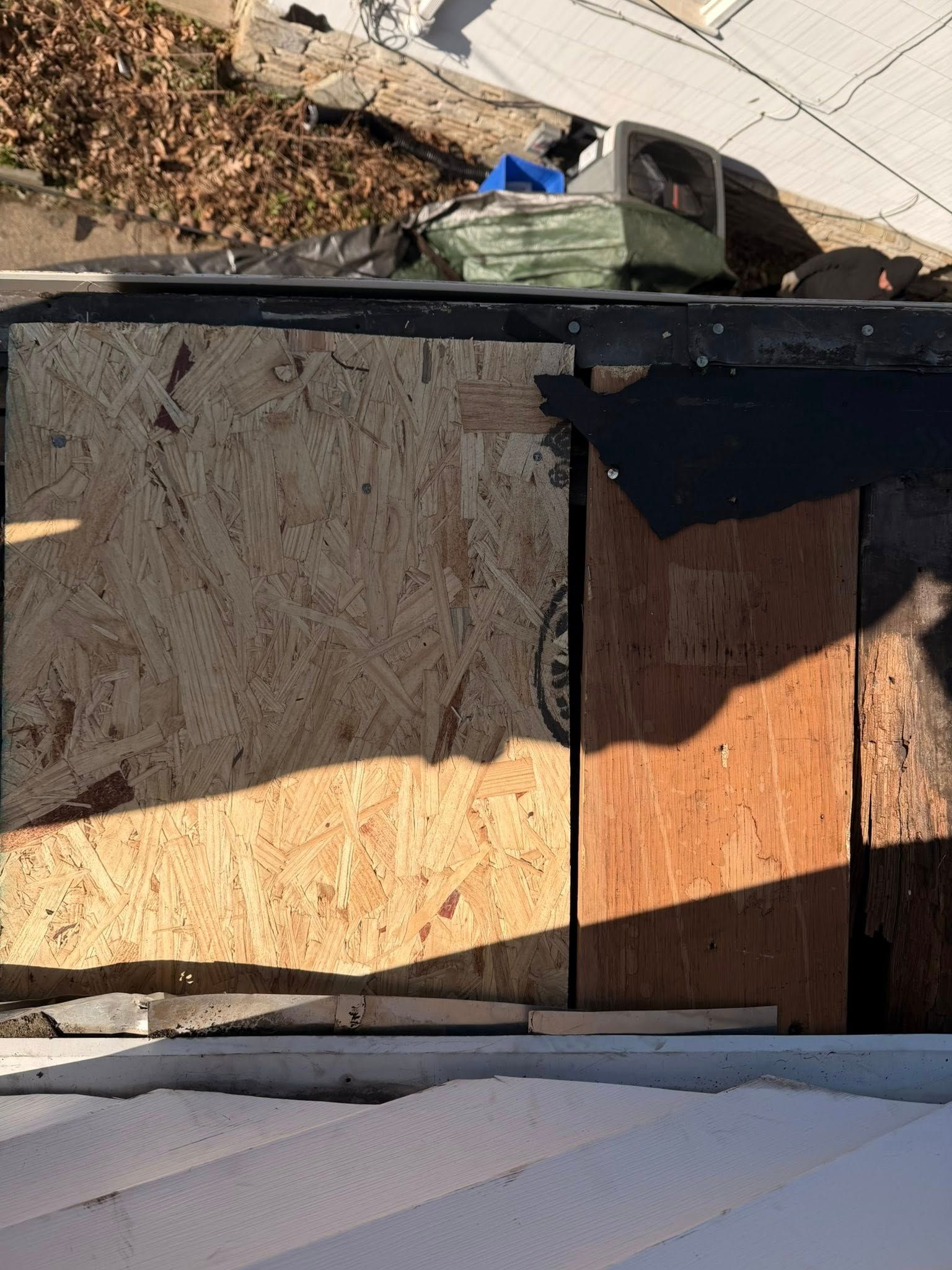 Two boarded-over windows on a damaged roof, with debris and a parked vehicle below.