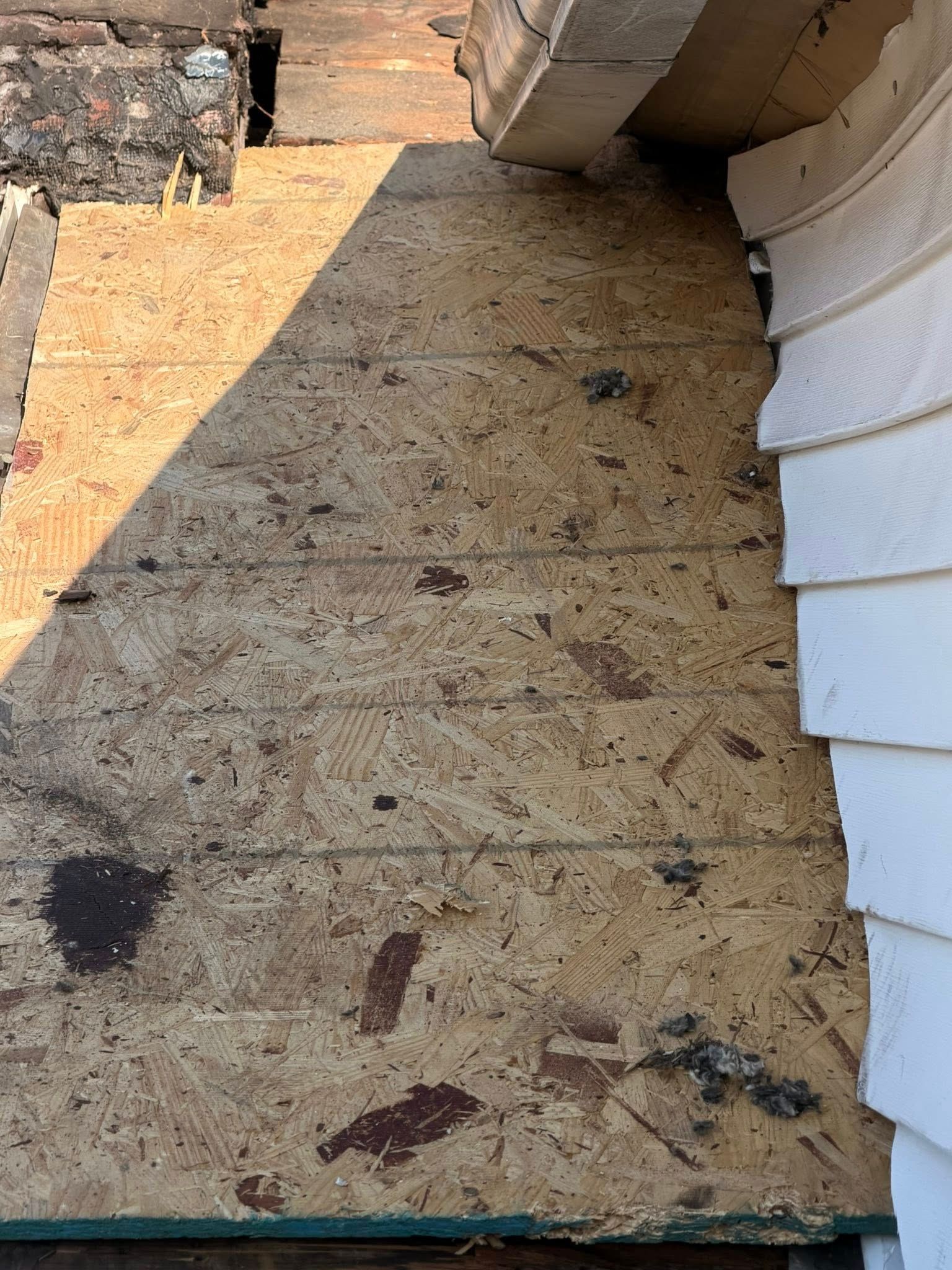 Weathered plywood rooftop walkway beside brick and white siding walls, with dark stains and debris.