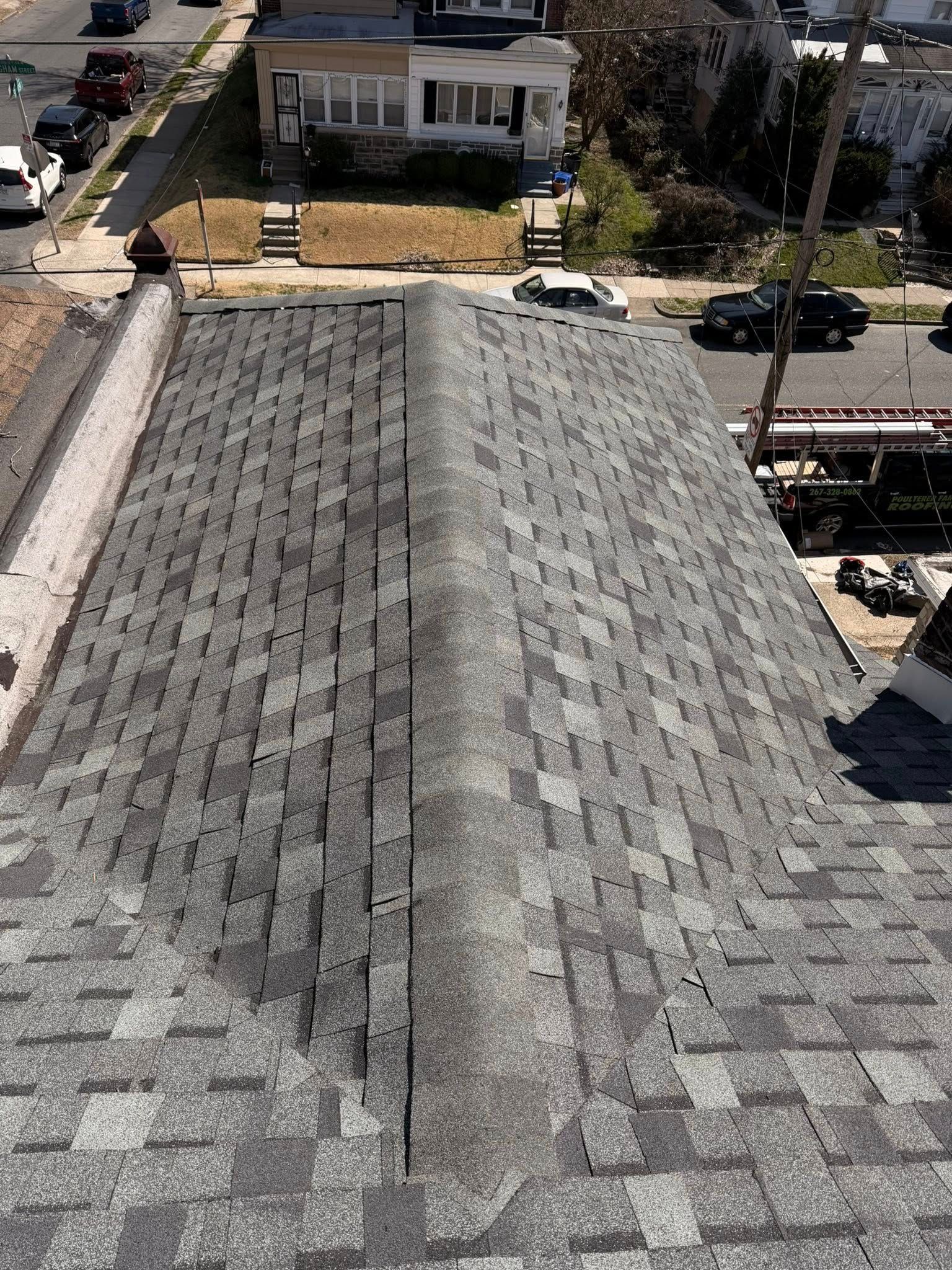 Aerial view of a shingled roof with a ridge vent, driveway, and houses along a street.