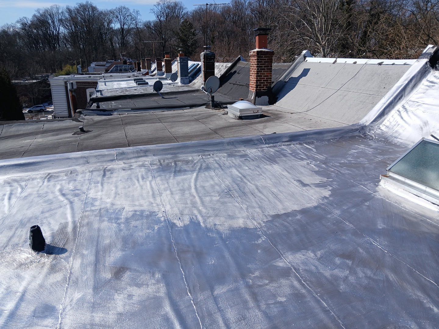 Flat rooftop with white waterproof coating, vents, and a brick chimney under a clear sky.