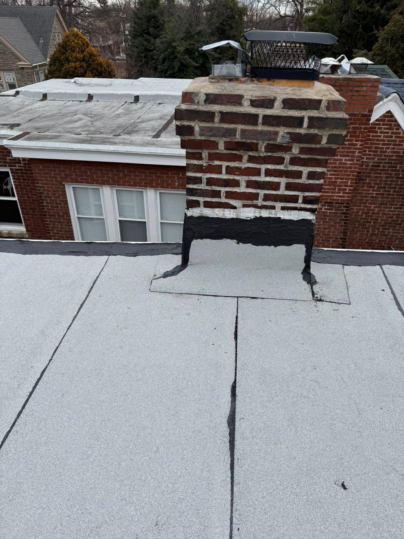 Rooftop view of a brick chimney with a black flashing patch on a gray flat roof