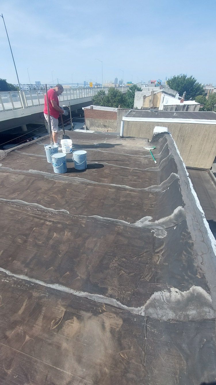 Rooftop with waterproofing work in progress; worker in red shirt near buckets and tools under a blue sky