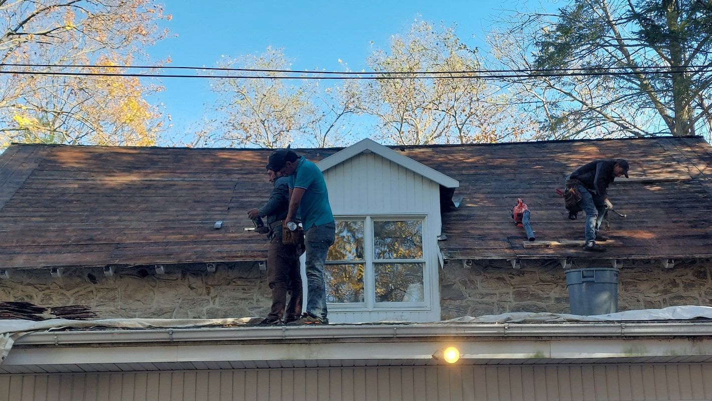 Two workers repair a shingled roof above a dormer window on a house.