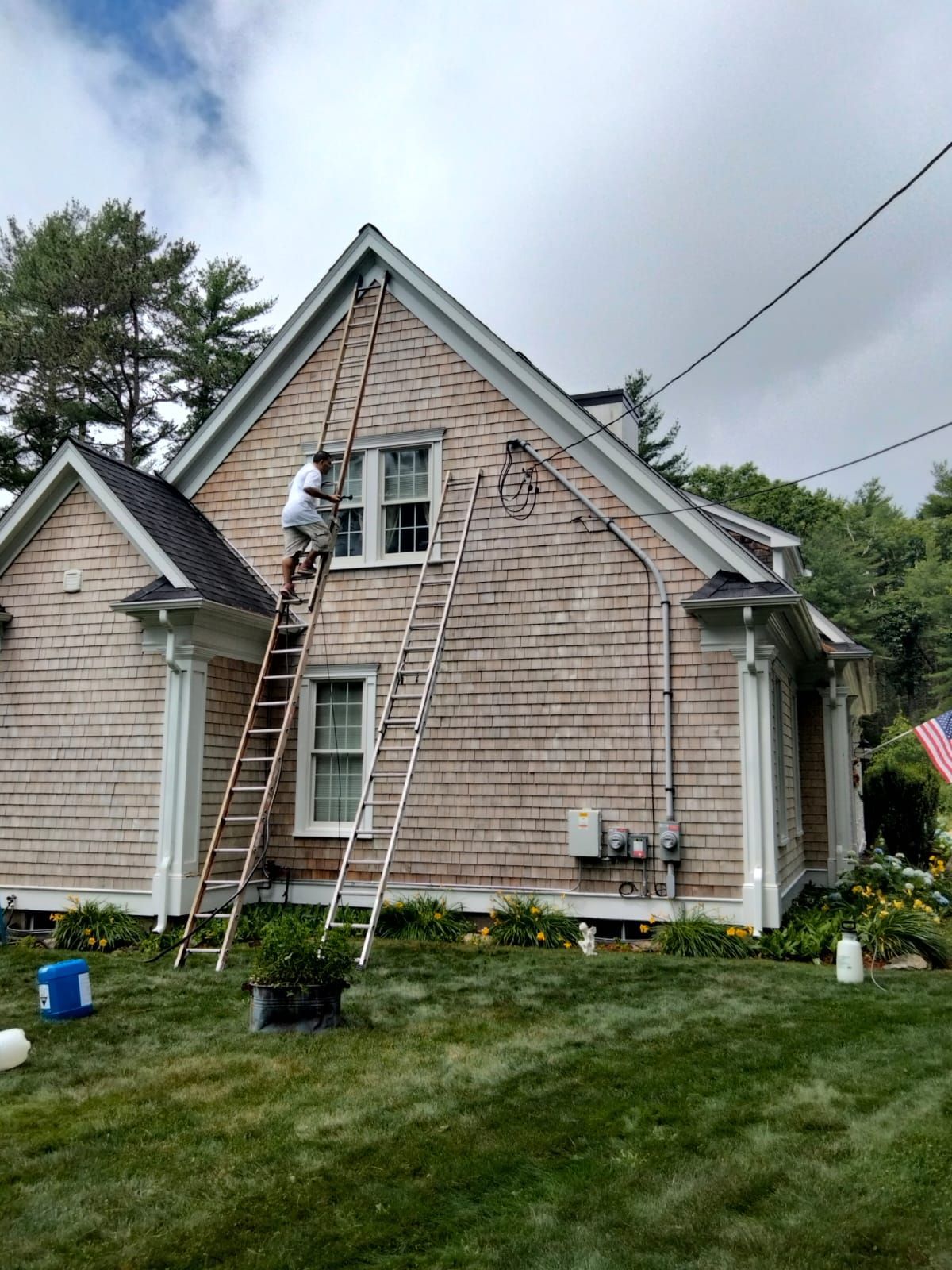 Person painting exterior of a house on a ladder, cedar shingles, cloudy day.