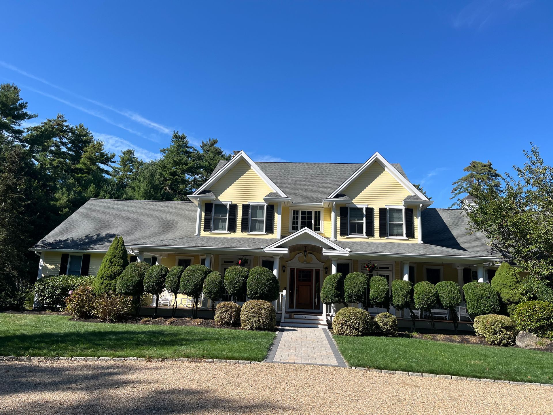 Yellow house with black shutters, shrubs, and a paved walkway under a blue sky.