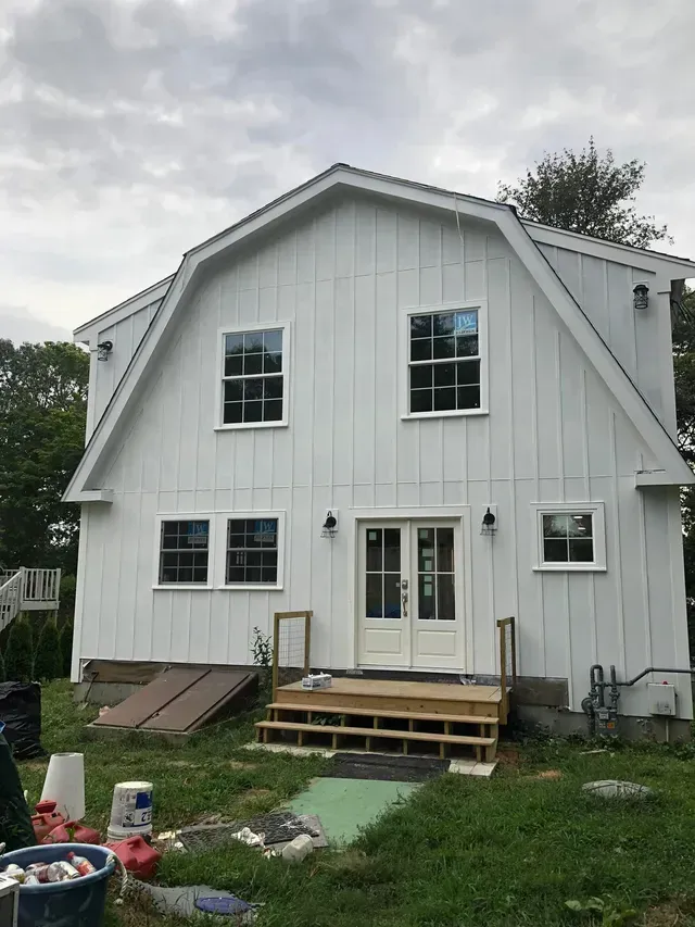 White barn-style house under construction, with windows, a double door, and unfinished steps on a cloudy day.