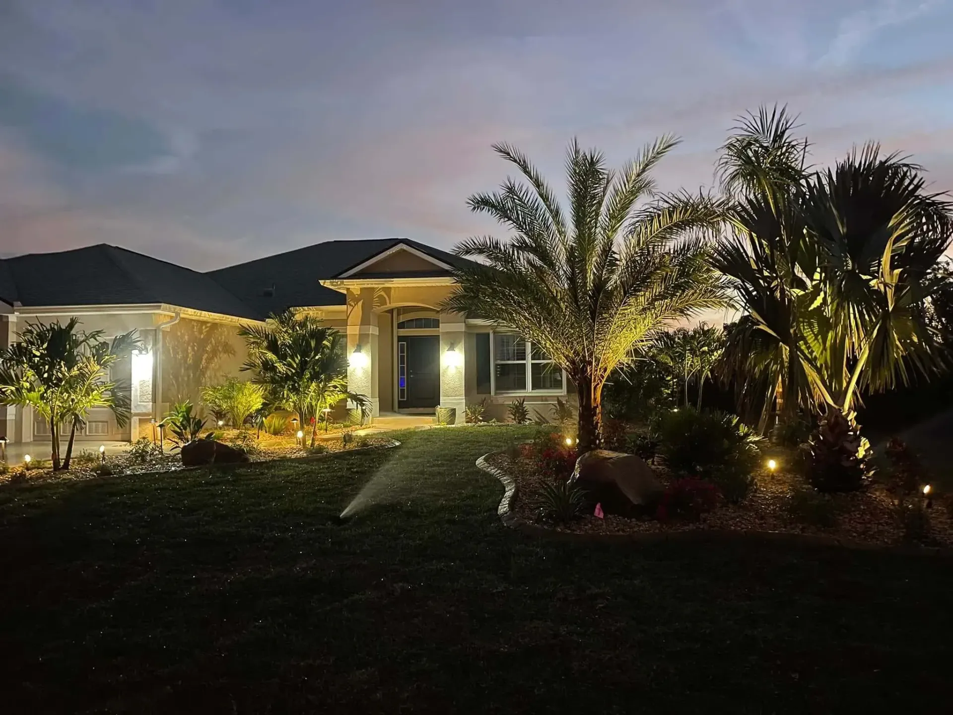 A lit modern house at dusk, with illuminated palm trees and landscaped garden in front.