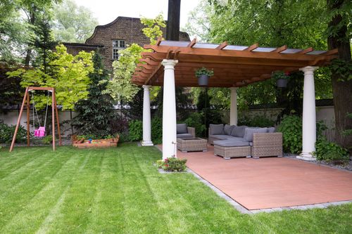 Backyard patio with pergola, wicker sofa, and swing set amid green lawn and trees