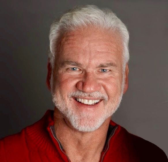 Smiling man with white hair and beard, wearing a red top, against a gray background.