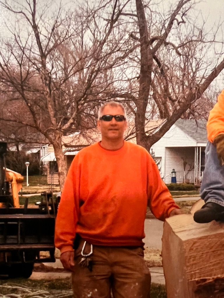 Man in orange sweatshirt and sunglasses standing outdoors beside a brick planter and trees